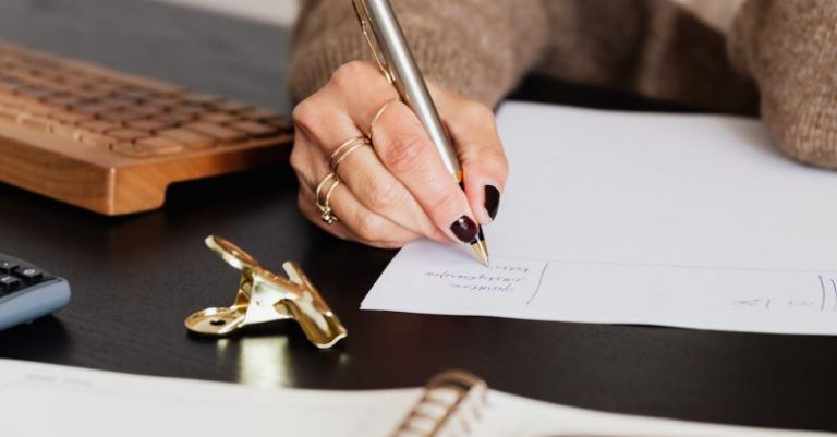 Smart Investments - Unrecognizable female entrepreneur writing on piece of paper while sitting at table with notebook and smartphone