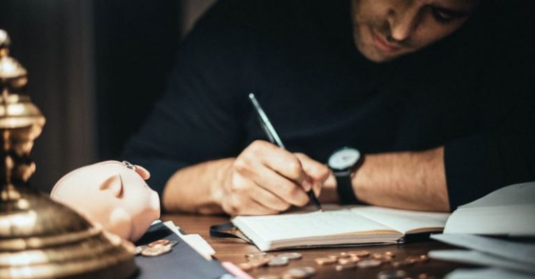 Smart Investments - Crop elegant man taking notes in journal while working at desk with coins and piggybank in lamplight
