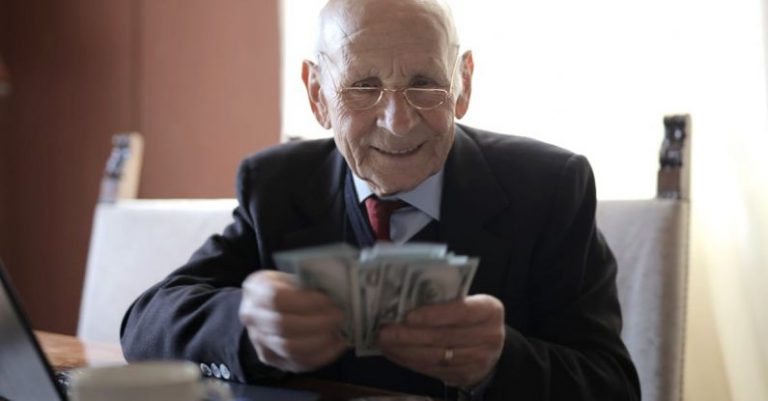Smart Investments - Positive senior businessman in formal suit and eyeglasses counting money bills while sitting at wooden table with cup of beverage and near opened laptop